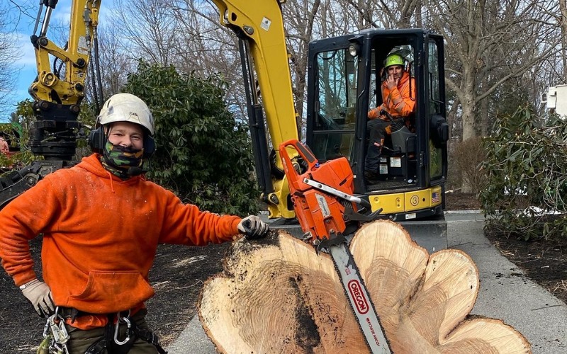 A Top Notch Tree Service crew member in safety gear posing with a chainsaw next to cut tree logs with excavator equipment nearby.