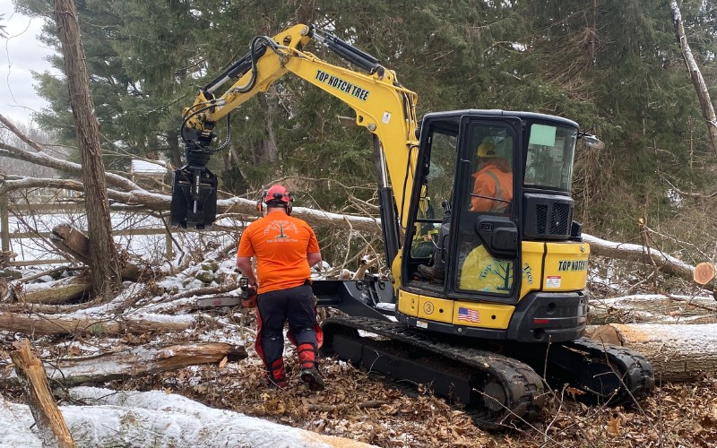 Two Top Notch Tree crew members in bright orange safety gear are working with a yellow excavator to clear fallen trees and debris in snowy winter conditions.