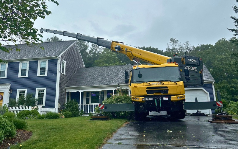 A large crane truck is positioned in a residential driveway for safe tree removal near a Massachusetts home.