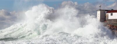 Large waves crashing against rocks near a white lighthouse during a coastal storm.